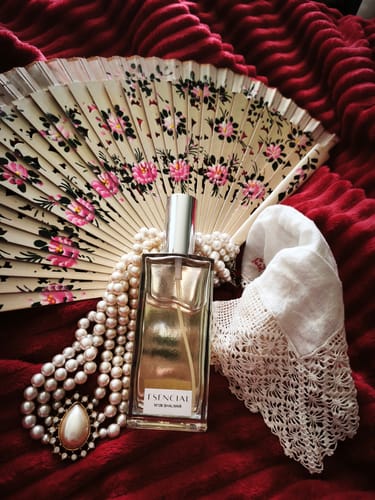 Un cliente muestra el perfume M106 en una elegante composición con un abanico, perlas y encaje sobre una tela roja.