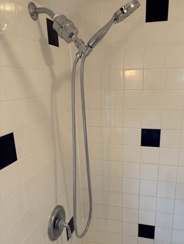 Reviewer's installed chrome handheld shower head and filter set against a white tiled shower wall.