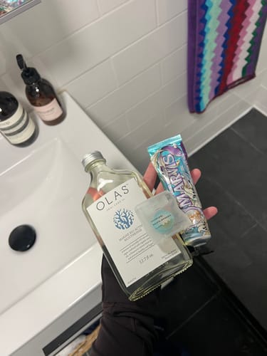 Customer holding a bottle of OLAS Mouthrinse with other toiletries in their hand over a bathroom sink.