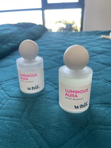 Reviewer's two bottles of Luminous Aura perfume, one new and one used, sitting on a bed.