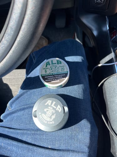 Customer's tin of Mountain Wintergreen Nicotine Pouches and a second branded grey tin resting on their lap in a car.
