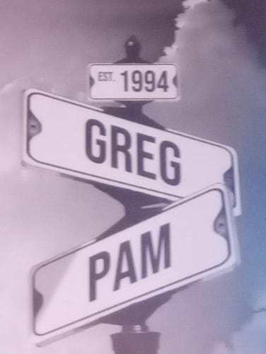 Customer's personalized 'Date of Love' canvas with street signs for 'Greg,' 'Pam,' and 'Est. 1994' against a cloudy sky.