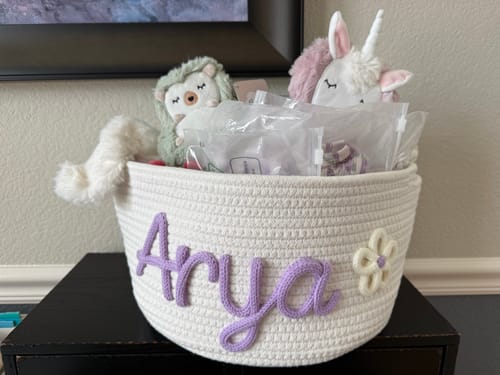 Customer's personalized white knitted basket with 'Arya' in purple yarn and a flower icon, holding baby shower gifts.