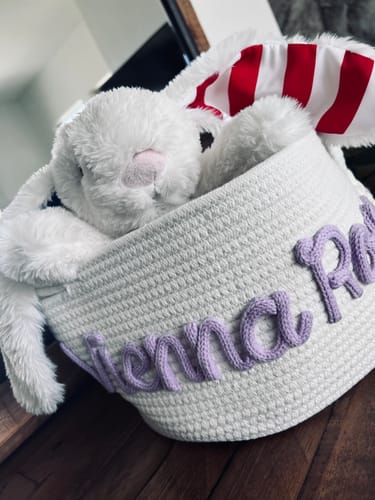 Close-up of a customer's white knitted basket with the name 'Yienna Ro...' in purple yarn, holding a stuffed bunny.