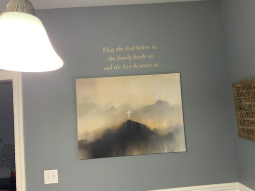 Customer's gold vinyl decal with the prayer 'Bless the food before us...' on a gray wall above a painting.