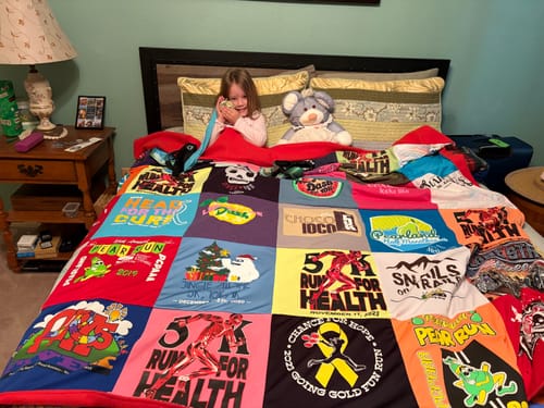 Customer's colorful Project Repat T-Shirt Quilt spread on a bed, with a child peeking out from underneath.