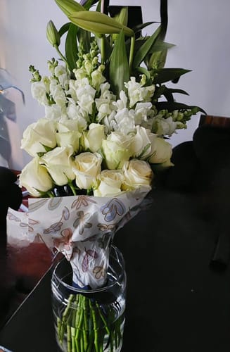 Customer's Pure Elegance Bouquet featuring fresh white roses and tall lilies in a clear glass vase on a dark surface.