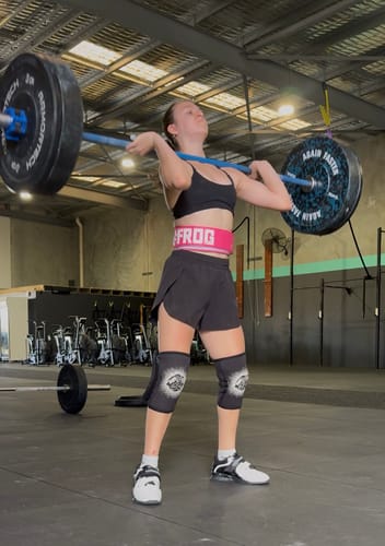 Reviewer wearing the bright Fluro Pink Lifting Belt while holding a barbell in a front rack position in a gym.