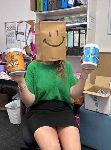 Reviewer playfully hiding their face with a bag, holding a tub of UGLYFACE® STIM and a tub of Hydroprime in an office.