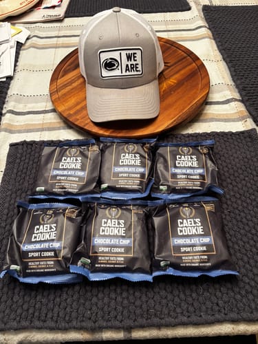 Customer review image showing six packages of Cael's Cookie Chocolate Chip arranged on a table with a 'We Are' hat.