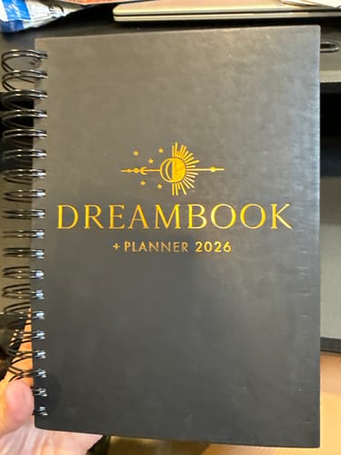 Customer holding the black 2026 Dreambook + Planner, showing its gold foil title and spiral binding.