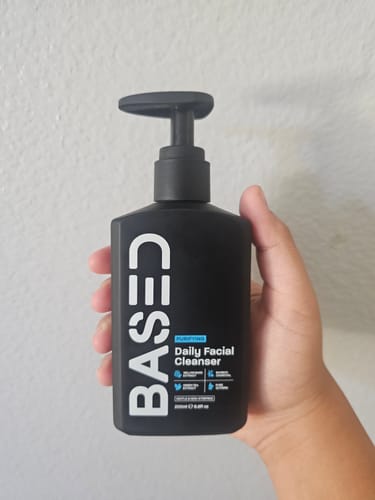 Reviewer holding the black pump bottle of BASED Daily Facial Cleanser against a white wall.