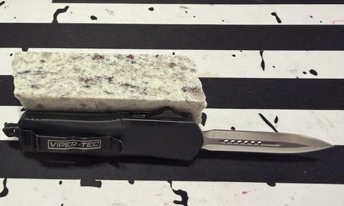 Reviewer's Small Black Ghost D/A OTF knife with its sharp blade extended on a striped surface.
