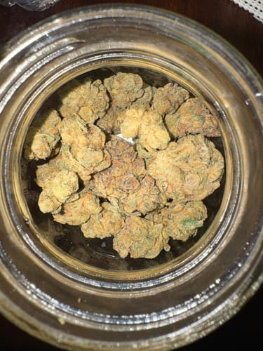 Reviewer's close-up of green and orange Jealousy THCa buds inside a clear glass jar.