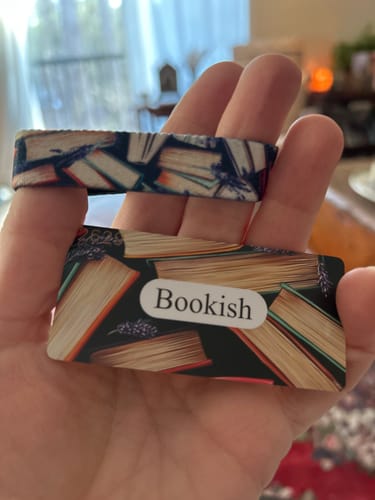 Reviewer's hand holding the Bookish Bracelet and its matching collector's card, which both show a colorful book pattern.