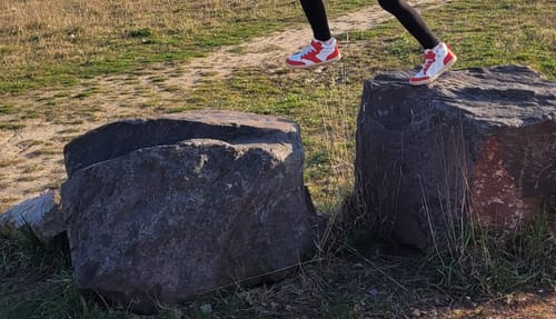 Ein Kind trägt die weiß-roten Kinder Wonderland I Lite Barfußschuhe beim Springen über Felsen im Freien.
