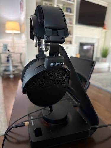Reviewer's black HEDDphone D1 headphones resting on a headphone stand in a living room setting.