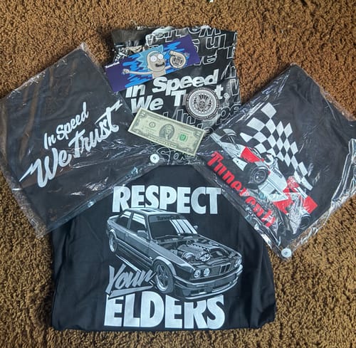 Customer's TCG92 MYSTERY CASH BOX contents, showing three graphic T-shirts, a sticker, and a two-dollar bill.