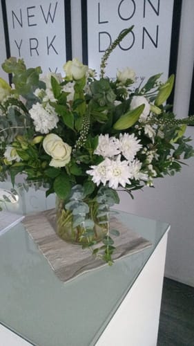 Customer's White Purity Vase Arrangement showing a large, lush bouquet of white flowers and greenery in a clear vase on a table.