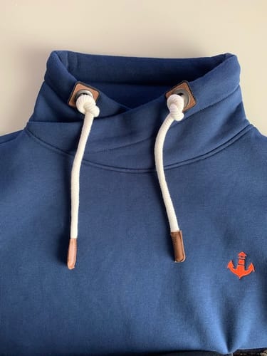 Kunde zeigt eine Nahaufnahme des blauen Schalkragen-Sweatshirts mit verknoteten weißen Kordeln und orangefarbenem Anker-Logo.
