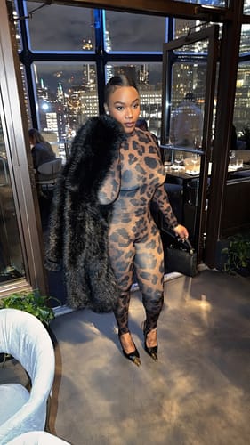 Customer posing in the Naked Print Stirrup Jumpsuit in a restaurant, holding a black fur coat.