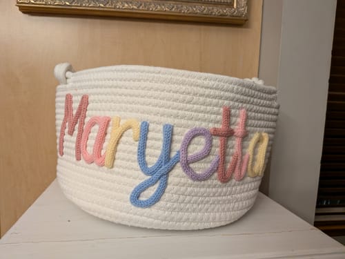 Customer's white knitted basket personalized with the name 'Maryetta' in colorful yarn on a white table.