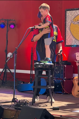 A customer's son wearing the custom black and red-royal baseball jersey on a stage with an adult.