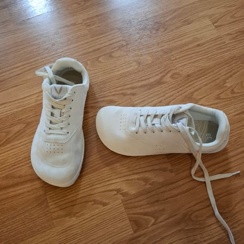 Asiakkaan valkoiset Vaistoa Katu 2 -paljasjalkasneakerit puulattialla.