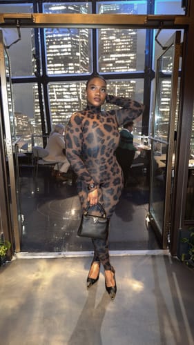 Customer in a full-length view of the Naked Print Stirrup Jumpsuit, standing in front of a city view window.