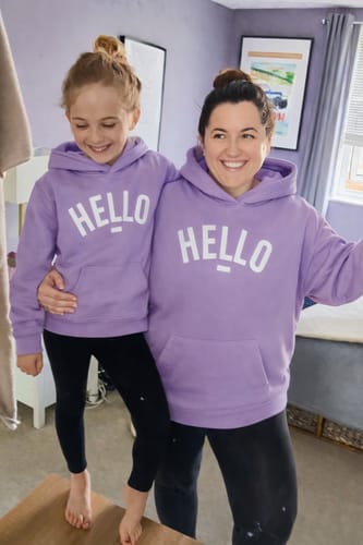 A reviewer and a child smiling while wearing matching lavender Hello Cowl Neck Hoodies indoors.