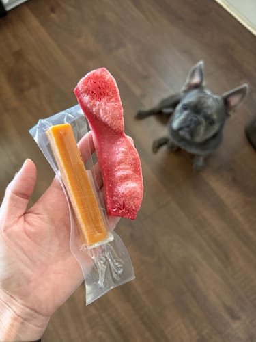 Customer holds an original Long-Lasting Yak Chew and a puffed red one, with a dog in the background.
