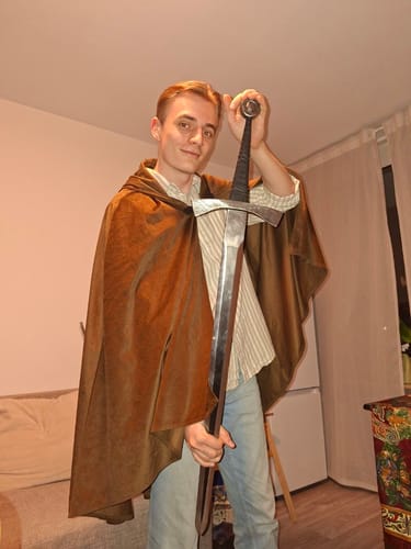 Reviewer in a brown cloak proudly holding the 'Wolf's Fury' steel sword upright indoors.