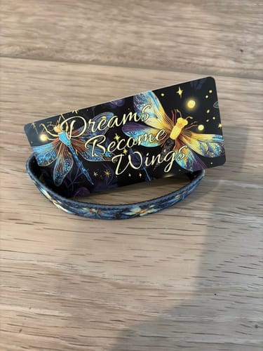 Reviewer's Dreams Become Wings Bracelet and its packaging card with dragonfly art, resting on a wooden surface.