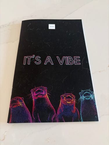 Customer's March 2026 Booklet featuring a black cover with four colorful otters and text that reads 'IT'S A VIBE'.