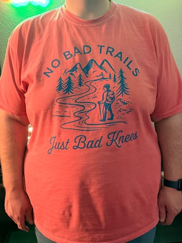 A reviewer wearing the coral-colored Bad Knees T-shirt, showing the relaxed fit on their torso.