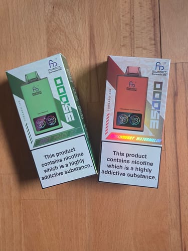 Ein Kunde zeigt zwei neue Verpackungen des RANDM TORNADO 35000 in Grün und Orange auf einer Holzoberfläche.