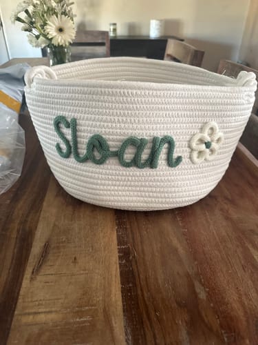 Customer's personalized white knitted basket with the name 'Sloan' in green yarn and a flower icon, sitting on a wooden table.