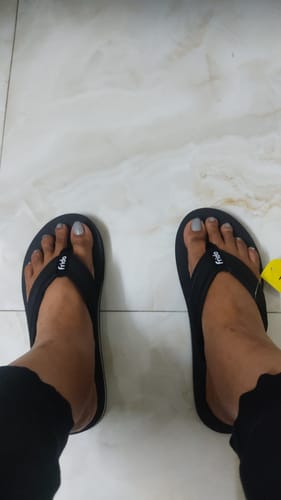 A reviewer's first-person view of their feet wearing the black Frido Cloud Comfort Arch Support Slippers on a tiled floor.