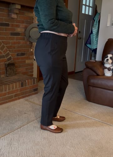 Reviewer showing the fit of the black Nicole Ankle Pants from the side while standing in a living room.
