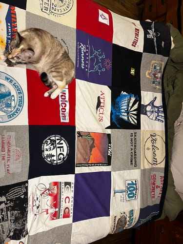 Reviewer's colorful Project Repat T-Shirt Quilt spread across a bed, with a cat resting on the corner.