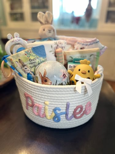 Customer's white personalized knitted basket with the name 'Paislee' in multicolor yarn, filled with assorted baby gifts.