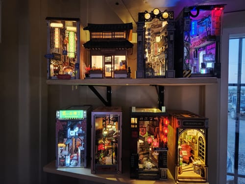 Reviewer's collection of lit book nook kits on two shelves, including the Kowloon Walled City 2049 kit.