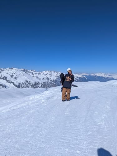 Reviewer standing on a snowy mountain wearing the baggy caramel-colored Snow Pants and holding a snowboard.