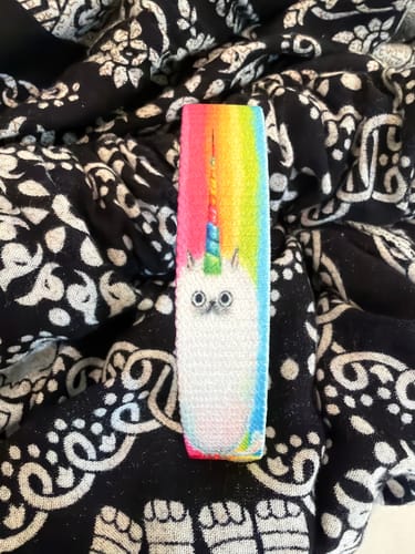 Reviewer's Expect Magic Bracelet with a caticorn design and rainbow background, shown flat on a patterned fabric.