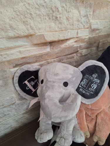 Ein Kunde zeigt einen grauen, personalisierten Plüschelefanten mit dem Namen und den Geburtsdaten des Babys auf den Ohren.