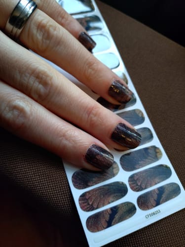 Eine Kundin zeigt ihre fertige Maniküre mit dunklen, gold-schimmernden 'On New Year's Wings' Nagelfolien.