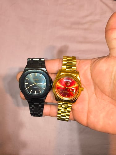 Reviewer's hand holding a gold Lucky Fours® Santos watch with a red face next to a black watch.