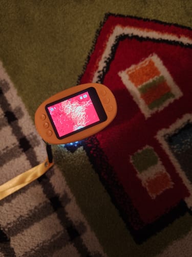 Ein Kunde zeigt das orangefarbene Abenteuer-Mikroskop, das die roten Fasern eines Teppichs vergrößert darstellt.