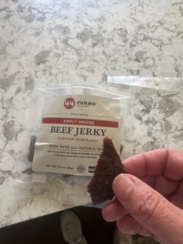 Customer's hand holding a piece of Simply Smoked beef jerky with the open package on a counter.