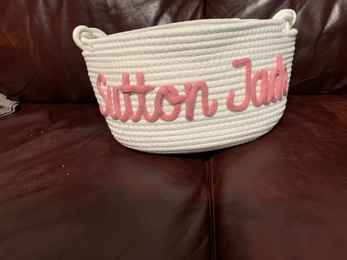 Customer's white knitted basket personalized with the name 'Sutton Jade' in pink yarn, sitting on a leather couch.